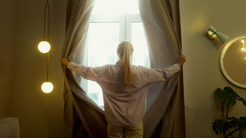 A woman stands by a window, opening curtains in a softly lit room.